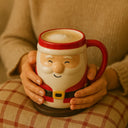 Pessoa segurando a caneca de Natal em cerâmica Papai Noel com as duas mãos, apoiada em almofada xadrez, transmitindo conforto.