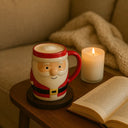 Caneca de Natal em cerâmica Papai Noel sobre suporte de madeira, ao lado de uma vela acesa e de um livro aberto, em cenário aconchegante.