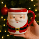 Caneca de Natal em cerâmica Papai Noel sendo segurada em frente a uma árvore de Natal iluminada e desfocada ao fundo.