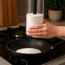 Mão utilizando tapioqueira preta com peneira embutida para polvilhar tapioca diretamente na frigideira – preparo limpo e eficiente.