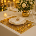 Mesa posta sofisticada com jogo americano dourado vazado, prato branco e arranjo floral iluminado – Casa Splendida.