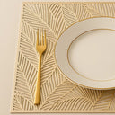 Jogo americano dourado retangular com detalhe vazado, composto com prato branco em mesa elegante.