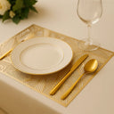 Composição refinada com jogo americano dourado, prato branco e talheres dourados em mesa posta moderna.