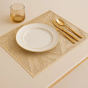 Jogo americano retangular dourado com textura vazada em mesa posta minimalista com prato e taça transparentes.