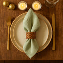 Anel de guardanapo em rattan claro rústico aplicado em guardanapo verde sobre prato claro, composição natural e sofisticada