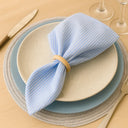 Anel de guardanapo em madeira natural aplicado em guardanapo azul claro sobre prato branco, composição clean e sofisticada