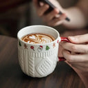 Close-up de caneca de cerâmica branca com textura de tricô e borda com símbolos de Natal, alça vermelha, cheia de chocolate quente com marshmallows