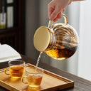 Bule de vidro transparente com tampa dourada e infusor interno, ideal para chás e infusões, sobre mesa de madeira