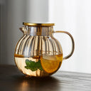 Bule de vidro transparente com tampa dourada e infusor interno, ideal para chás e infusões, sobre mesa de madeira