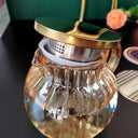 Bule de vidro transparente com tampa dourada e infusor interno, ideal para chás e infusões, sobre mesa de madeira